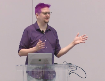 Screenshot of Adam in purple hair, gesturing dynamically, while presenting this talk.